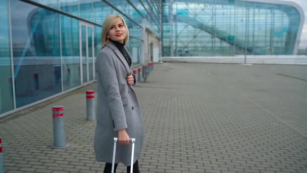Blond meisje rolt een koffer in de buurt van de luchthaven terminal - uitzicht vanaf de achterkant. Slow motion — Stockvideo