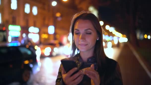 Donna attraente utilizza uno smartphone mentre cammina per le strade della città sera — Video Stock