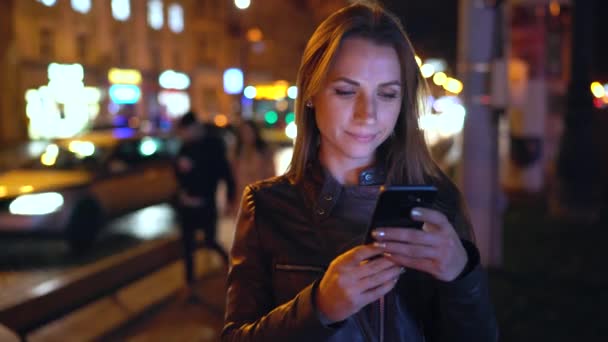 Приваблива жінка використовує смартфон під час прогулянки вулицями вечірнього міста — стокове відео