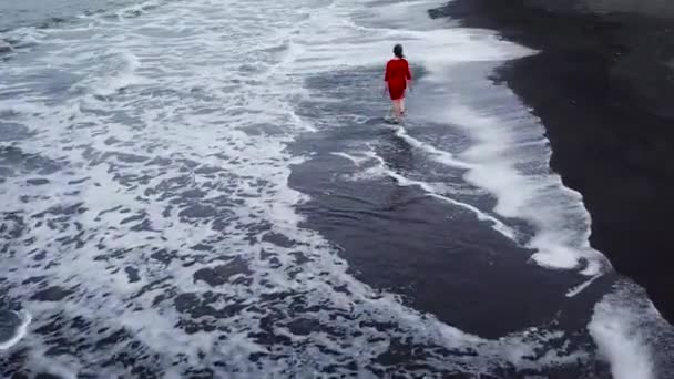 Αεροφωτογραφία του ένα κορίτσι σε ένα κόκκινο φόρεμα, περπάτημα στην παραλία με μαύρη άμμο. Τενερίφη, Κανάριοι Νήσοι, Ισπανία — Αρχείο Βίντεο