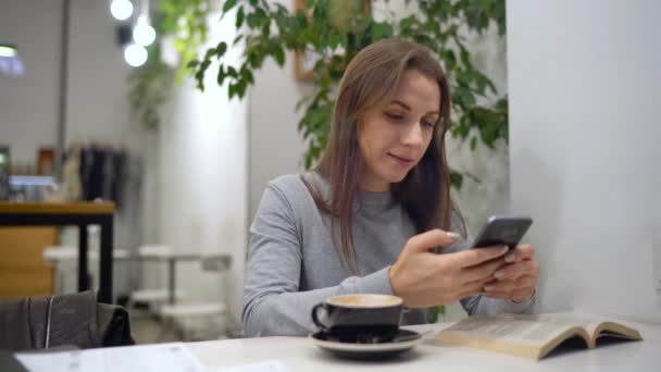 Красива жінка проводить час в кафе: використовує смартфон і п'є каву — стокове відео