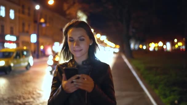 Ελκυστική γυναίκα χρησιμοποιώντας ένα smartphone, ενώ το περπάτημα στους δρόμους της πόλης το βράδυ — Αρχείο Βίντεο