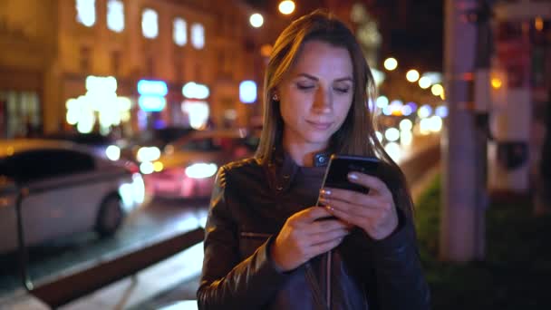 Mujer atractiva usando un teléfono inteligente mientras camina por las calles de la ciudad nocturna — Vídeos de Stock