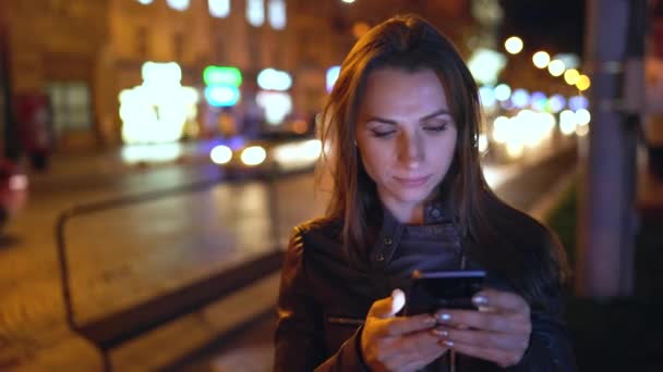 Aantrekkelijke vrouw met behulp van een smartphone tijdens het wandelen door de straten van de stad 's avonds — Stockvideo