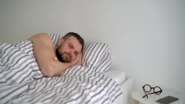 Hombre barbudo despertando en la cama debajo de la manta, sonriendo, poniéndose las gafas y tomando su teléfono inteligente — Vídeo de stock
