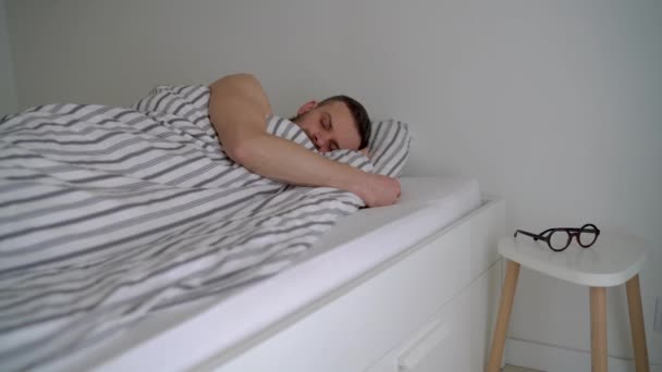 Bärtiger Mann wacht unter Decke im Bett auf, lächelt, setzt seine Brille auf und klatscht — Stockvideo