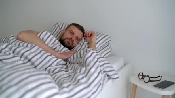 L'uomo barbuto si sveglia nel letto sotto la coperta, sorride, indossa gli occhiali e prende il suo smartphone — Video Stock