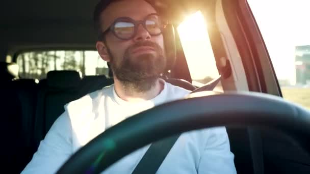 Hombre en gafas está estacionado en un coche en un estacionamiento al aire libre al atardecer — Vídeo de stock