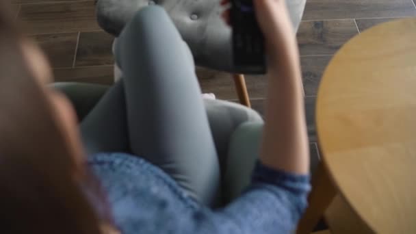 La mujer está sentada en una silla, viendo la televisión con una pantalla verde, cambiando los canales con un control remoto. Clave de croma. En interiores — Vídeos de Stock