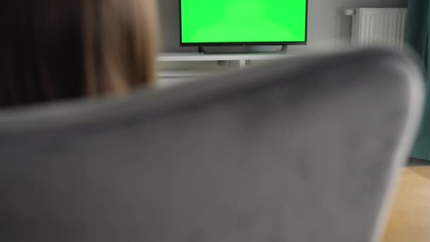 La mujer está sentada en una silla, viendo la televisión con una pantalla verde, cambiando los canales con un control remoto. Clave de croma. En interiores — Vídeo de stock