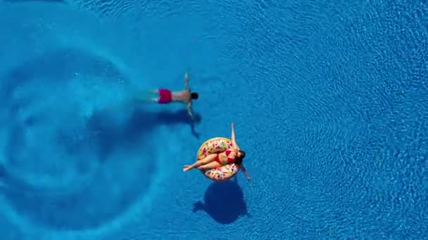 Vista aérea del hombre se sumerge en la piscina mientras la chica está acostada en una piscina de donuts flotar — Vídeos de Stock