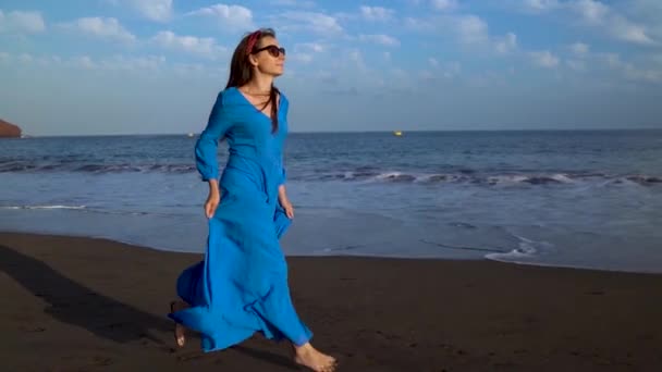 Kobieta w niebieskiej sukni biegnie wzdłuż czarnej plaży wulkanicznej. Zwolnionym — Wideo stockowe