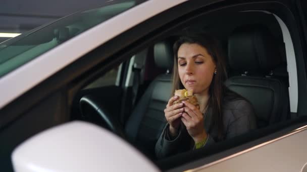 Женщина ест сэндвич, сидя в машине на парковке. Концепция современной насыщенной жизни — стоковое видео