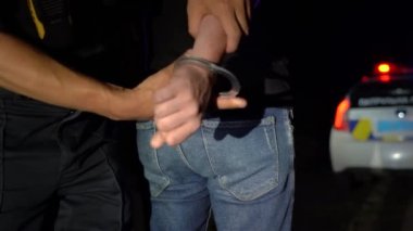 Polis memuru gece tutuklanan adama kelepçe takıyor ve onu polis arabasına götürüyor.