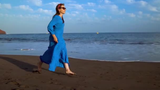 Mulher em um vestido azul corre ao longo de uma praia vulcânica preta — Vídeo de Stock