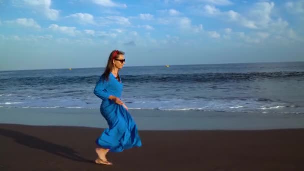 Kobieta w niebieskiej sukni biegnie wzdłuż czarnej wulkanicznej plaży — Wideo stockowe