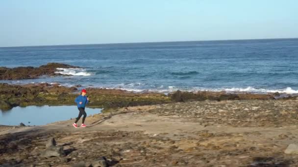 Una donna corre lungo la riva pietrosa dell'oceano. Stile di vita sano e attivo — Video Stock