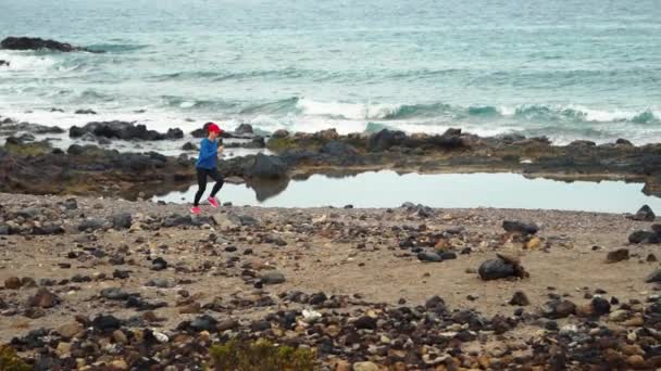 Una donna corre lungo la riva pietrosa dell'oceano. Stile di vita sano e attivo. Rallentatore — Video Stock