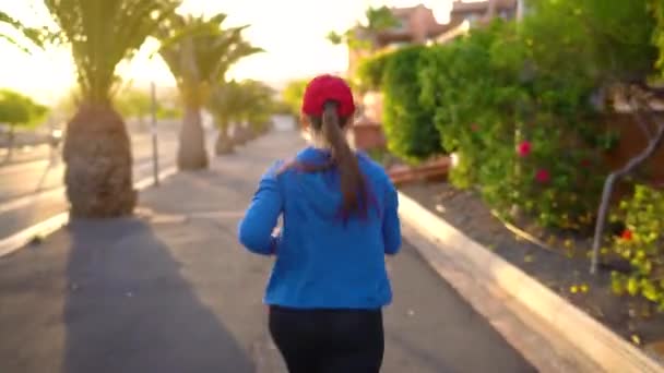 La mujer corre por la calle entre las palmeras al atardecer, vista trasera. Vida activa saludable — Vídeos de Stock