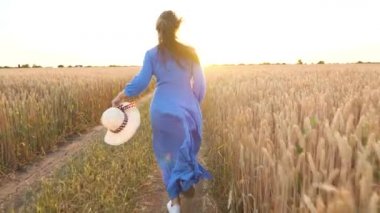 Mavi elbise li ve şapkalı güzel kadın gün batımında buğday tarlasında koşar. Özgürlük kavramı. Gün batımında buğday tarlası. Yavaş çekim