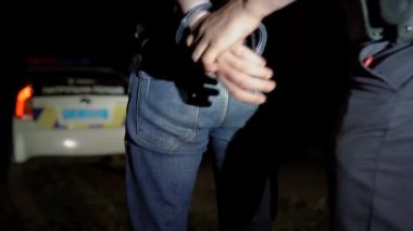 Polis memuru gece tutuklanan adama kelepçe takıyor ve onu polis arabasına götürüyor.