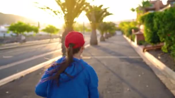 Frau läuft bei Sonnenuntergang die Straße hinunter zwischen den Palmen, Rückansicht. Gesunder aktiver Lebensstil — Stockvideo