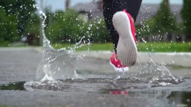 Jambes d'un coureur en baskets. Femme sportive faisant du jogging à l'extérieur, se jetant dans la flaque boueuse. Un seul coureur court sous la pluie, faisant des éclaboussures. Vue de derrière. Mouvement lent — Video