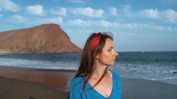 Frau mit Sonnenbrille und blauem Kleid, die an einem schwarzen Vulkanstrand spaziert. Zeitlupe — Stockvideo