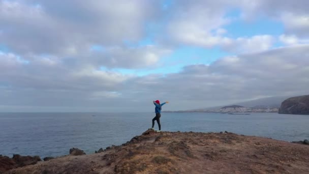 Luchtfoto van de vrouw in de sportkleding na het joggen staat op de top van een rots en geniet van het landschap in een beschermd gebied aan de oevers van de Atlantische Oceaan. Tenerife, Canarische eilanden, Spanje — Stockvideo