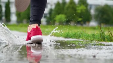 Spor ayakkabılı bir koşucunun bacakları. Spor kadını dışarıda koşuyor, çamurlu su birikintisine giriyor. Yağmurda koşan, su sıçratan tek koşucu. Geri görünüm
