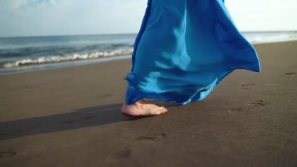 Pernas de uma mulher em belo vestido azul andando ao longo de uma praia vulcânica preta. Movimento lento — Vídeo de Stock
