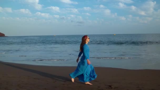 Femme en lunettes de soleil et robe bleue courant le long d'une plage volcanique noire — Video