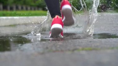 Spor ayakkabılı bir koşucunun bacakları. Spor kadını dışarıda koşuyor, çamurlu su birikintisine giriyor. Yağmurda koşan, su sıçratan tek koşucu. Arka görüş. Yavaş çekim