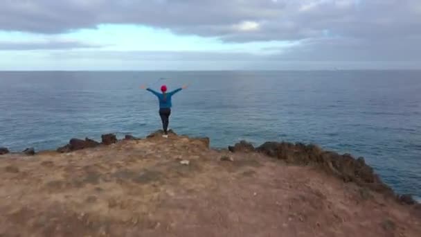 Vliegen over vrouw in sportkleding staat op de top van een rots met armen uitgestrekt aan de zijkanten en geniet van het landschap in een beschermd gebied aan de oevers van de Atlantische Oceaan. Tenerife, Canarische eilanden — Stockvideo