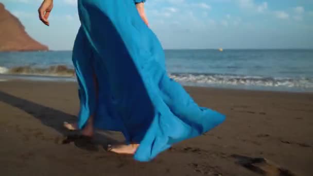 Beine einer Frau in wunderschönem blauen Kleid, die an einem schwarzen Vulkanstrand spaziert — Stockvideo