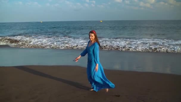 Mujer en un hermoso vestido azul y gafas de sol disfruta de un paseo por la playa, girando alegremente. Movimiento lento — Vídeos de Stock