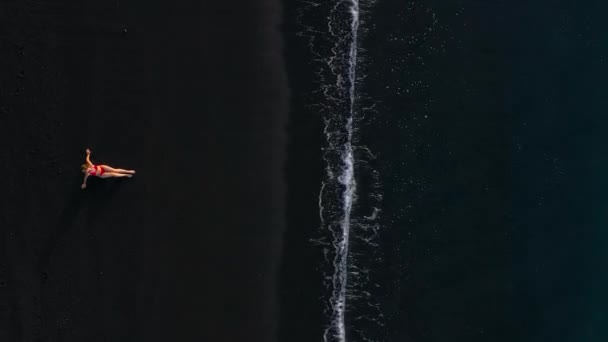 Vista superior de uma menina em um maiô vermelho deitado em uma praia preta na linha de surf. Costa da ilha de Tenerife, Ilhas Canárias, Espanha. — Vídeo de Stock