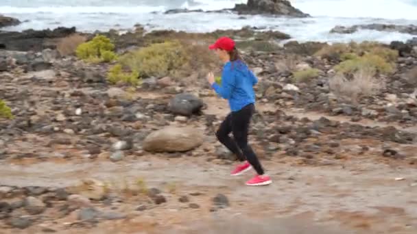Una donna corre lungo la riva pietrosa dell'oceano. Stile di vita sano e attivo — Video Stock