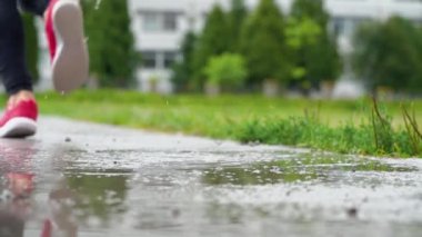 Spor ayakkabılı bir koşucunun bacakları. Spor kadını dışarıda koşuyor, çamurlu su birikintisine giriyor. Yağmurda koşan, su sıçratan tek koşucu. Arka görüş. Yavaş çekim