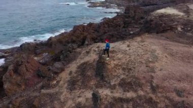 Koşu sonrası spor giyimli kadının havadan görünümü bir kayanın üzerinde duruyor ve Atlantik Okyanusu kıyılarındaki bir koruma alanında manzaranın keyfini çıkarıyor. Tenerife, Kanarya Adaları, İspanya