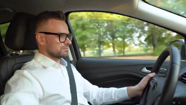 Homme barbu à lunettes et chemise blanche conduisant une voiture par temps ensoleillé. Vue latérale — Video