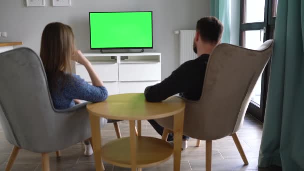 Mężczyzna i kobieta siedzą na krzeseł, oglądając telewizję z zielonym ekranem, dyskutują o tym, co zobaczyli i przełączają kanały za pomocą pilota zdalnego sterowania. Widok z tyłu. Klucz chrominancji. Pomieszczeniach — Wideo stockowe