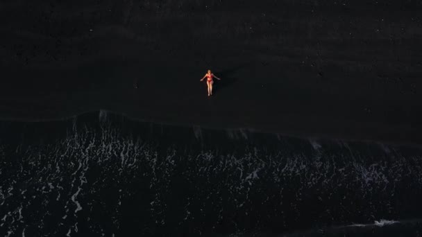 Top view of a girl in a red swimsuit lying on a black beach on the surf line. Coast of the island of Tenerife, Canary Islands, Spain. — Stock Video