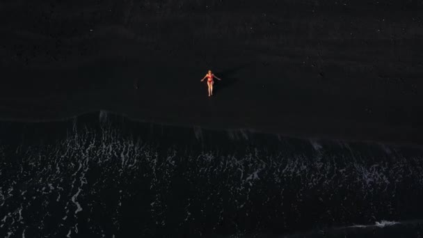 Vista superior de uma menina em um maiô vermelho deitado em uma praia preta na linha de surf. Costa da ilha de Tenerife, Ilhas Canárias, Espanha. — Vídeo de Stock