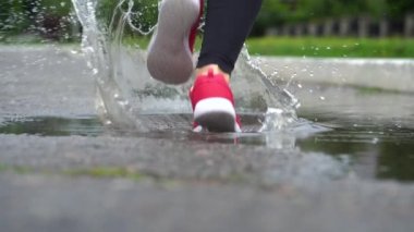Spor ayakkabılı bir koşucunun bacakları. Spor kadını dışarıda koşuyor, çamurlu su birikintisine giriyor. Yağmurda koşan, su sıçratan tek koşucu. Yavaş çekim