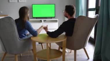 Erkek ve kadın sandalyelerde oturuyorlar, yeşil ekranla televizyon izliyorlar, uzaktan kumandayla kanal değiştiriyorlar. Geri görünüm. Renk anahtarı. Kapalı