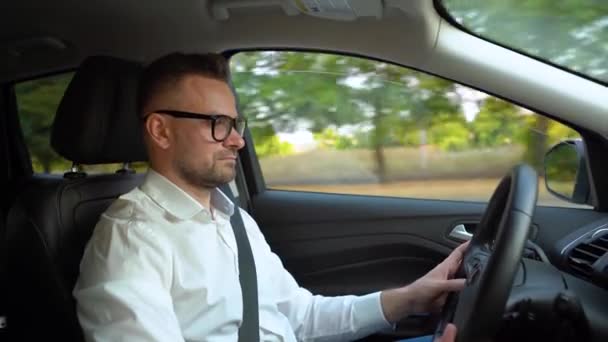 Bearded man in glasses and white shirt driving a car in sunny weather and uses autopilot function while driving — Stock Video