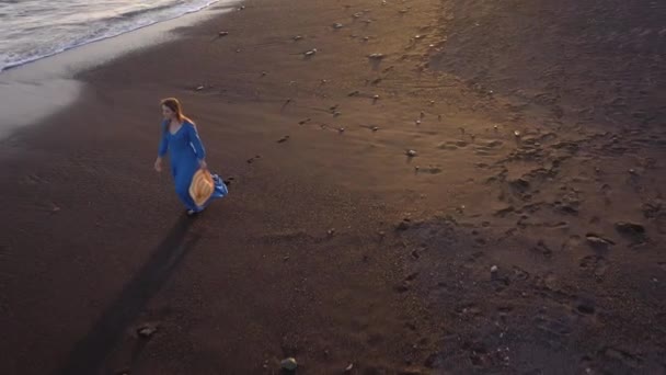 Vista aérea de una chica en un vestido azul caminando por la playa con arena negra al atardecer. Tenerife, Islas Canarias, España — Vídeo de stock