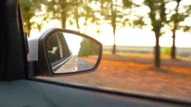 Gün batımında ağaçlarla çevrili yolda araba sürerken dikiz aynasından görünümü