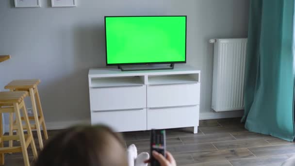 Frau sitzt auf einem Stuhl, schaut Fernsehen mit grünem Bildschirm und schaltet mit einer Fernbedienung die Kanäle. Chroma-Schlüssel. drinnen — Stockvideo